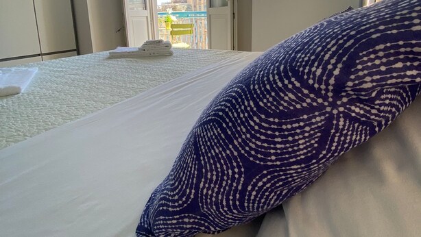 Une chambre avec un lit, un coussin à motifs, une vue sur la ville par une porte ouverte et un balcon avec une balustrade.