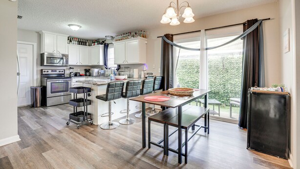 A modern kitchen with a dining area, featuring a large window with curtains, a chandelier, and a refrigerator.