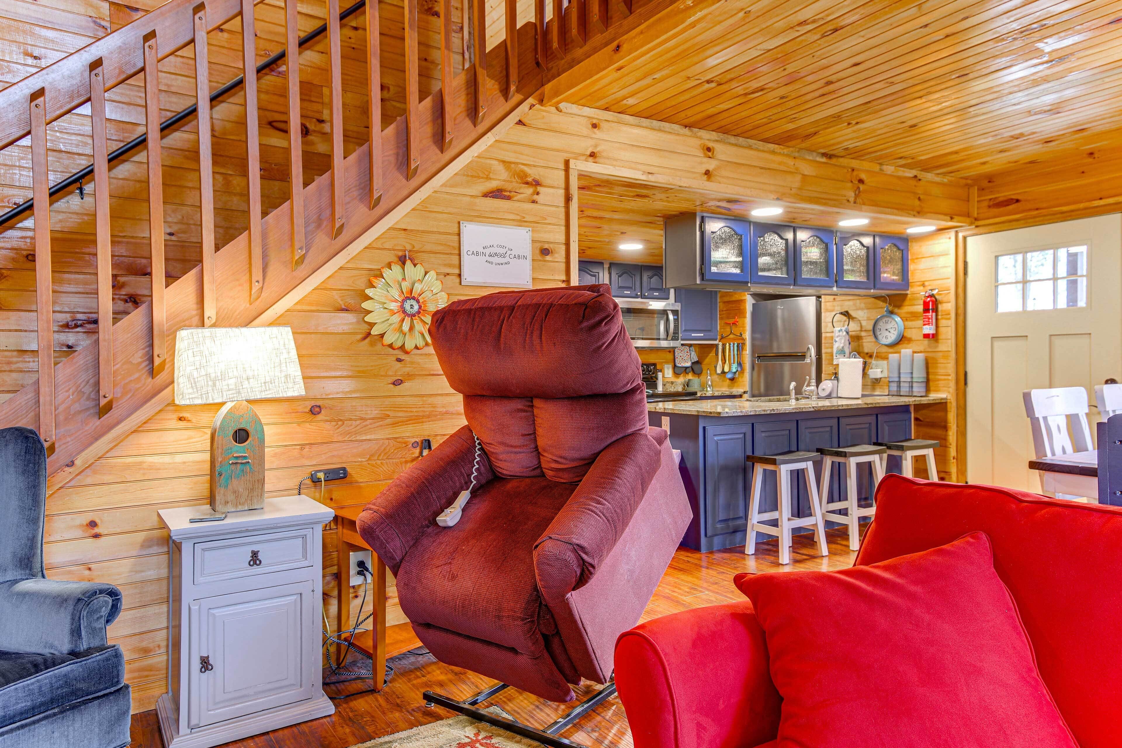 A cabin interior with a wooden staircase, a red recliner, a white cabinet, and a kitchen area with blue cabinets and a stainless steel refrigerator.