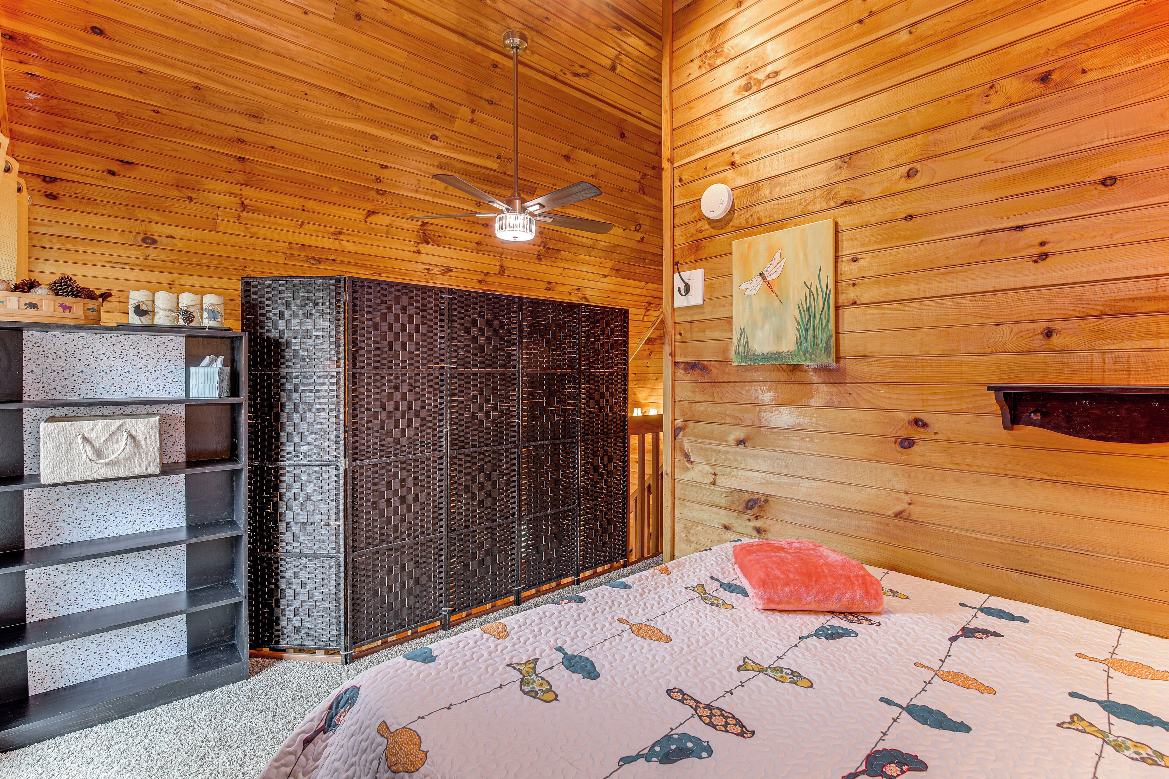 A wooden cabin interior with a bed, a ceiling fan, a shelf with decorative items, and a wall-mounted painting.