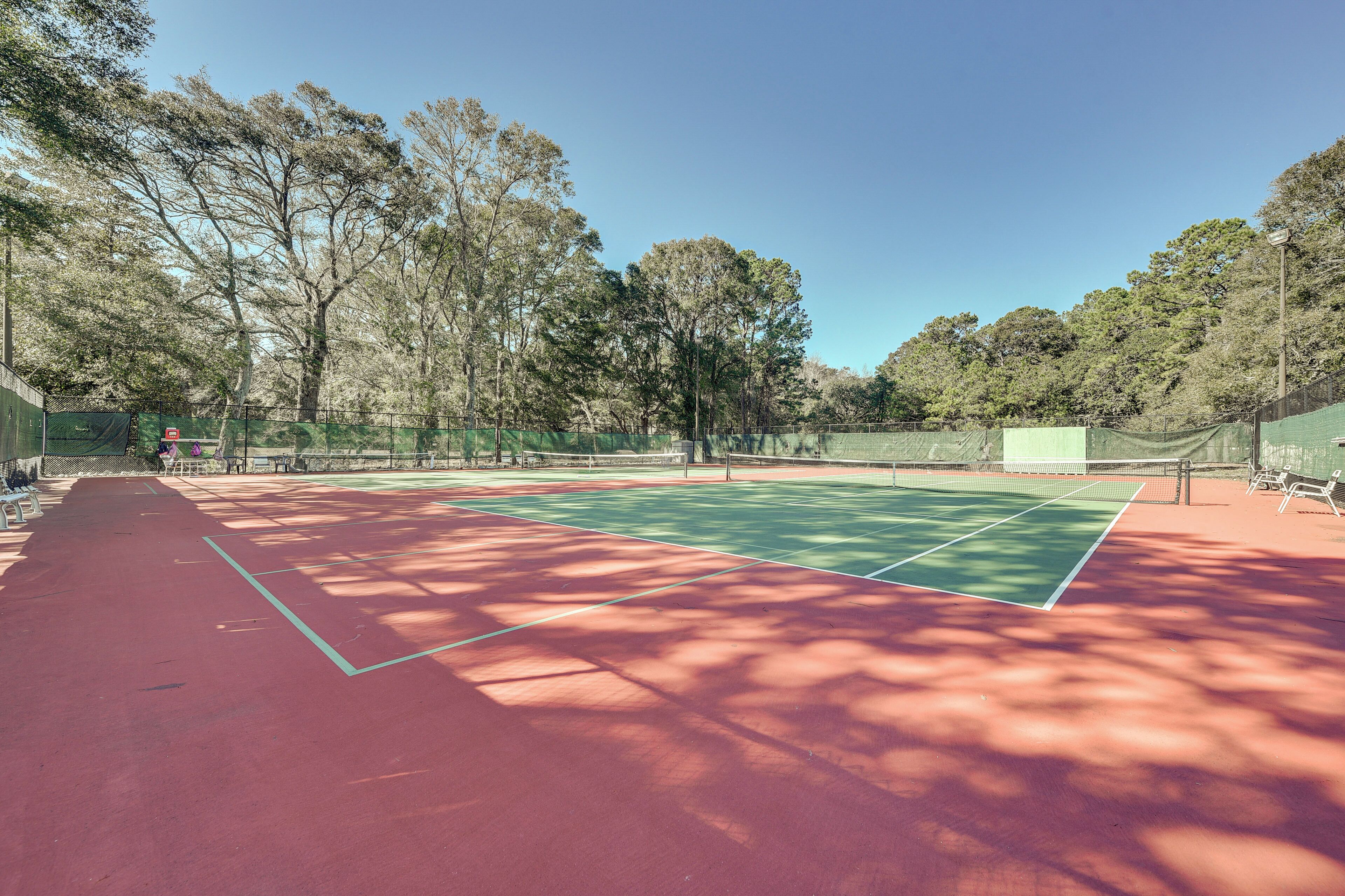 Ein Tennisplatz im Freien mit grün-roter Belag, umgeben von Bäumen und einem Zaun.