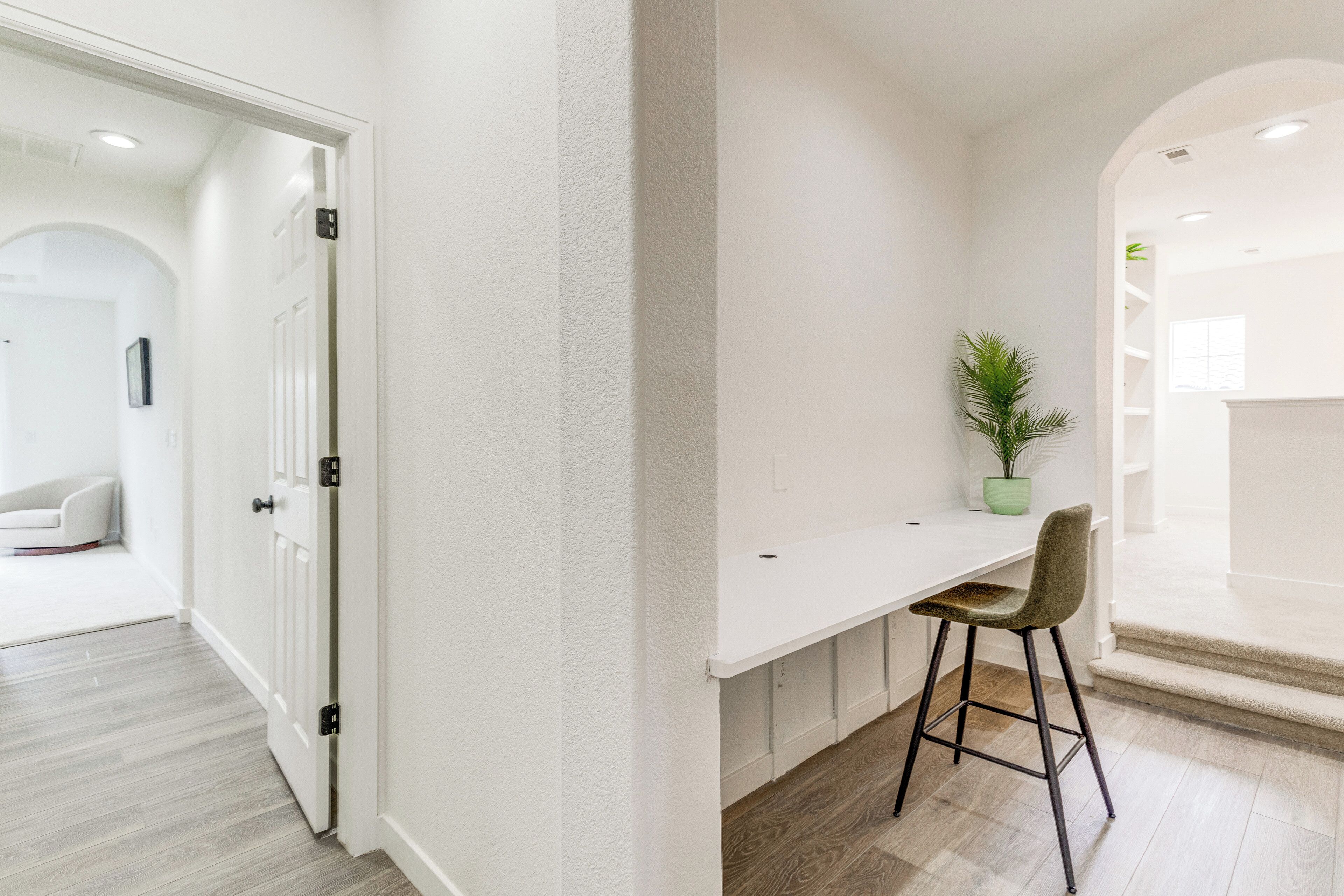 A modern, minimalist room with a white desk and chair, a potted plant, and a staircase leading to another area.