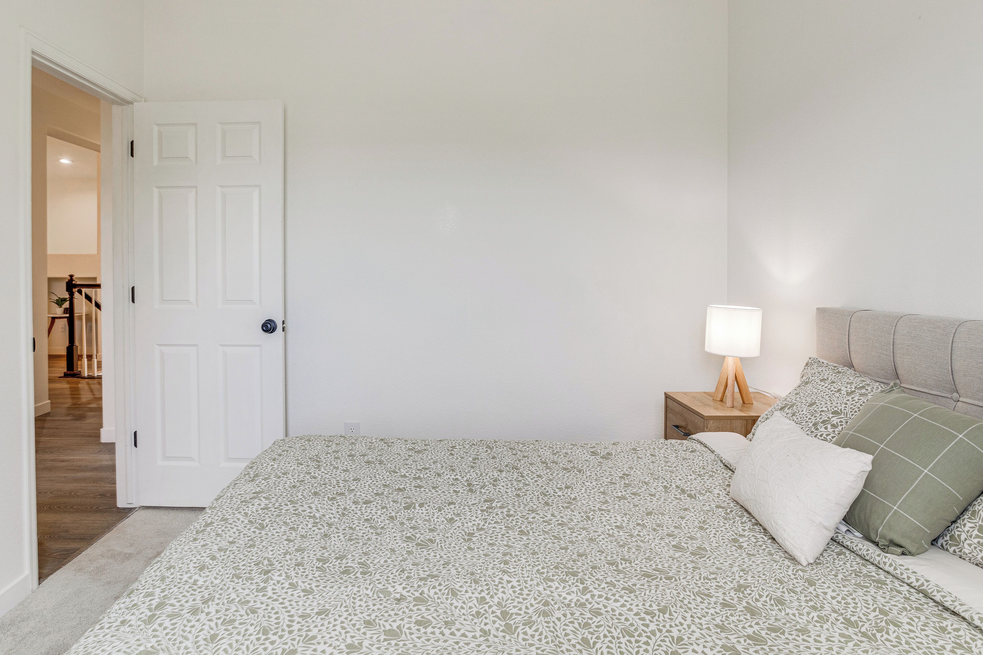 A bedroom with a bed, a nightstand with a lamp, and a door leading to another room.