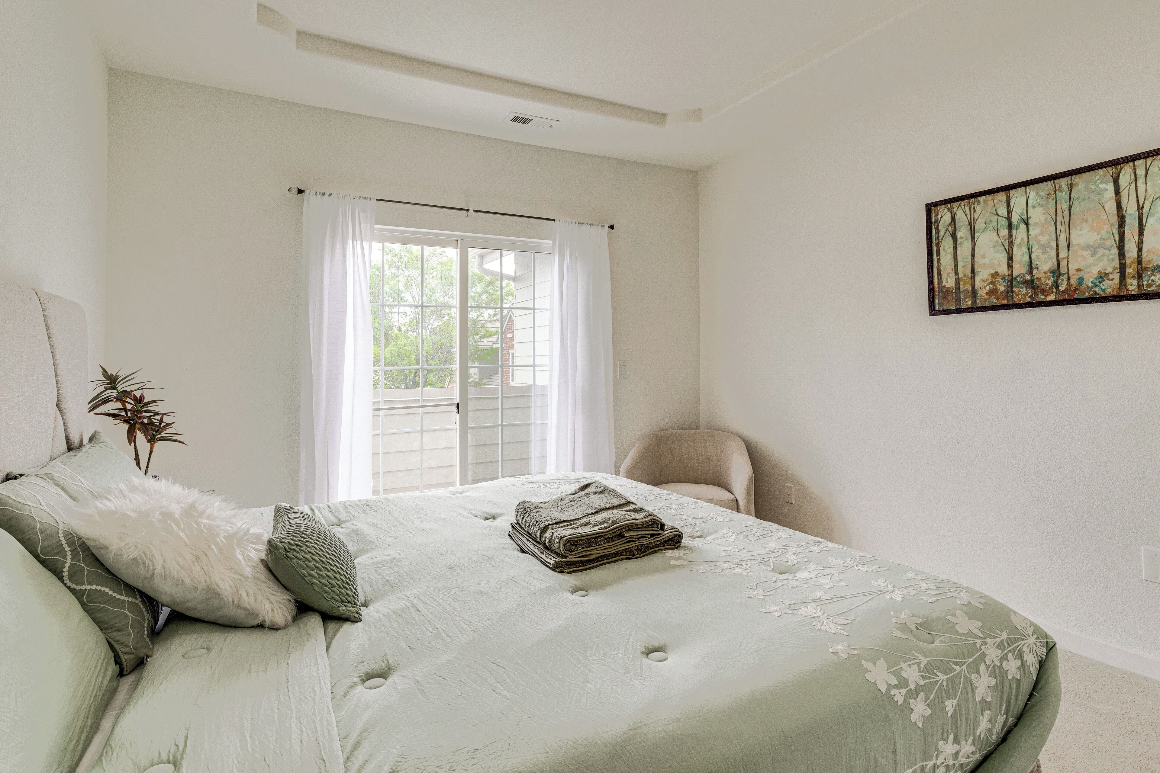 A bedroom with a bed, a chair, a window with curtains, and a painting on the wall.