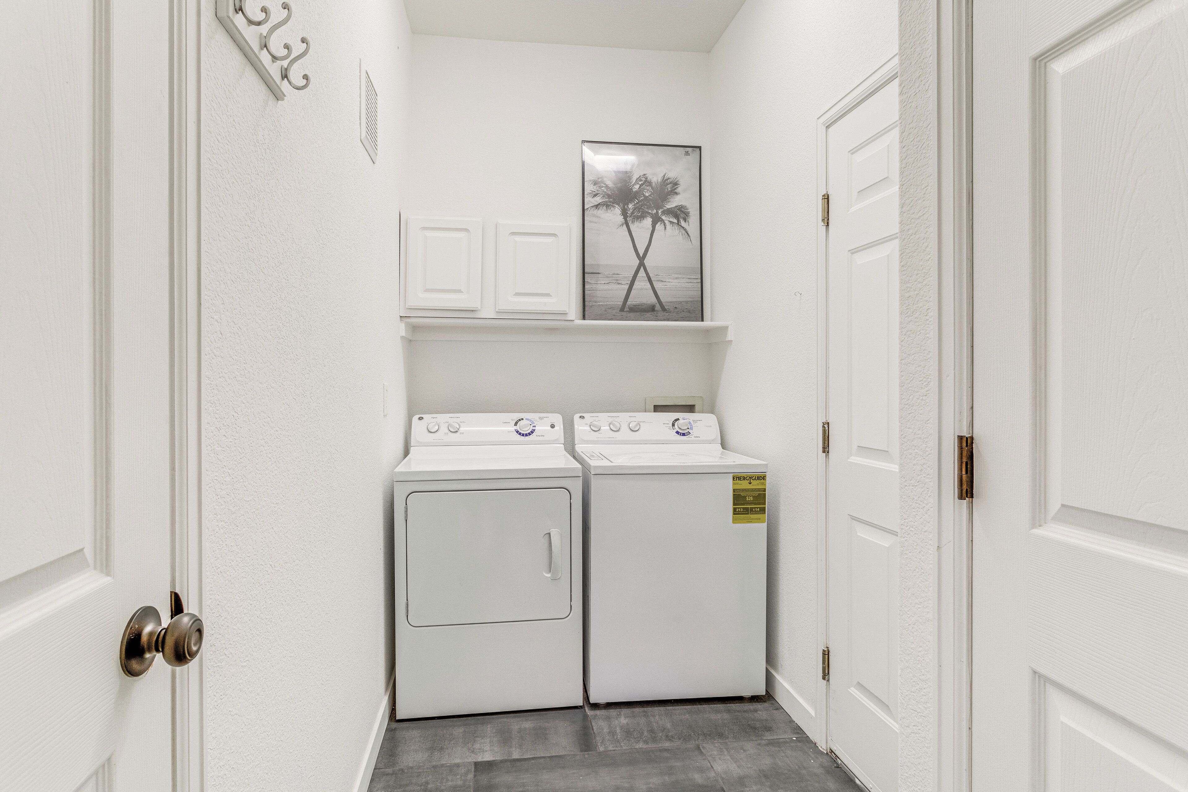 A laundry room with two white washers and dryers, a white door, and a framed picture of palm trees on the wall.
