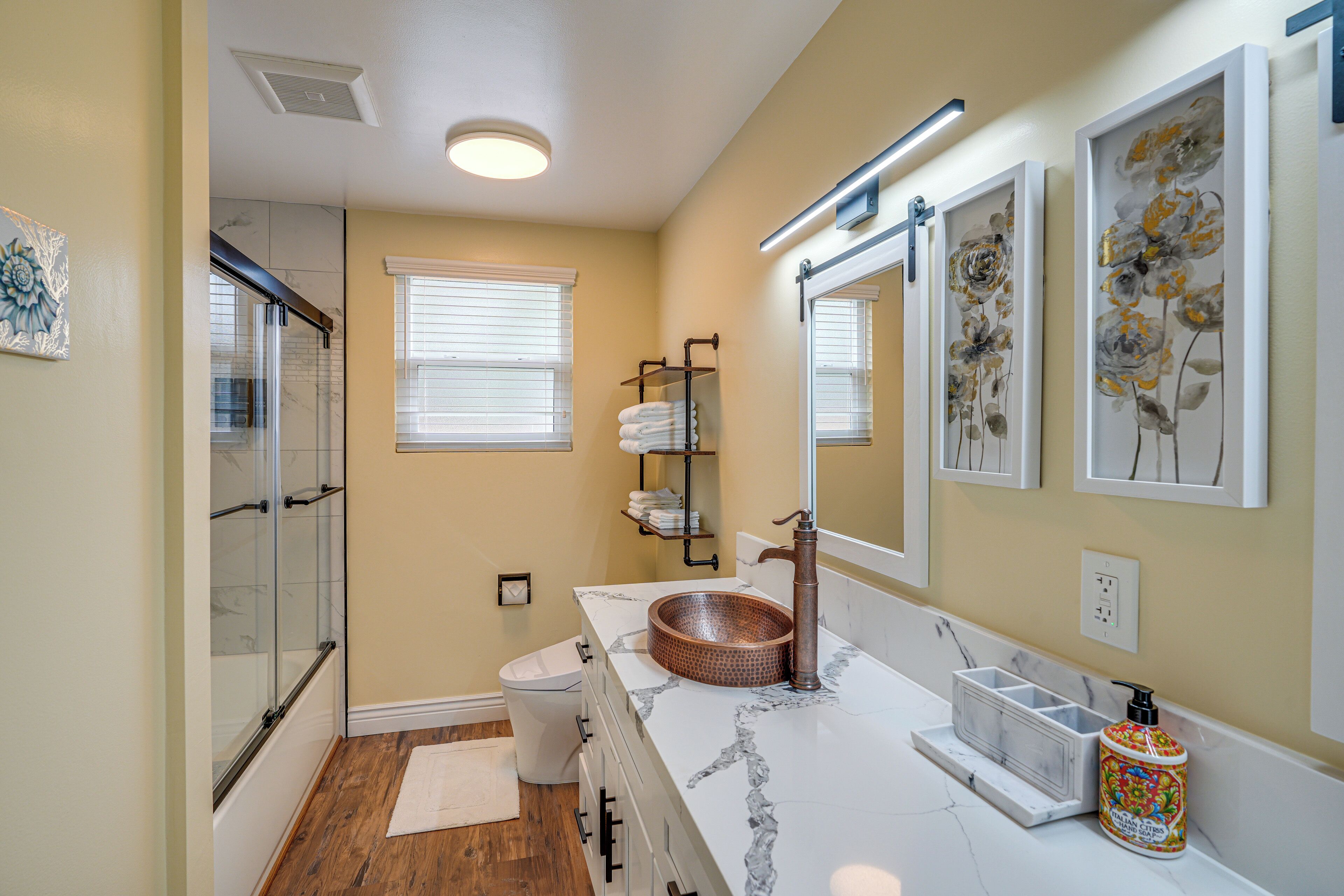 A bathroom with a glass shower enclosure, a white toilet, a marble sink with a copper basin, and a mirror with floral artwork.