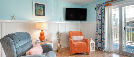 A living room with a blue wall, wooden flooring, a flat-screen TV, two armchairs, a small table with a lamp, and a window with curtains.
