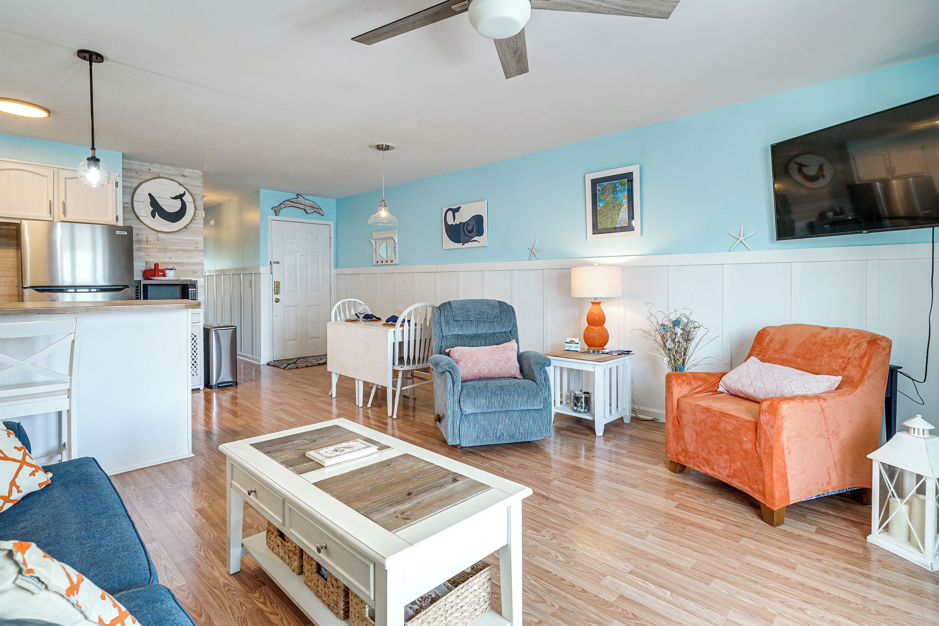 A modern living room with a kitchen in the background, featuring a blue wall, white furniture, and a ceiling fan.