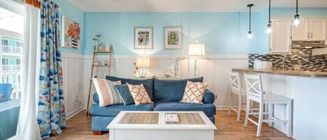 A living room with a blue sofa, white coffee table, and a kitchen area with white cabinets and a bar counter.