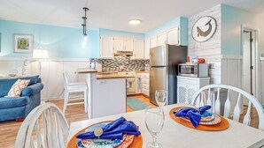 A modern kitchen with white cabinets, a stainless steel refrigerator, and a microwave. There is a dining area with a table set for two, featuring a glass and a plate with a blue napkin. - Carolina Beach Condo w/ Balcony: Walk to the Ocean (Carolina Beach)