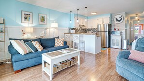A modern living room with a blue sofa, white coffee table, and a kitchen area with white cabinets and stainless steel appliances. - Carolina Beach Condo w/ Balcony: Walk to the Ocean (Carolina Beach)
