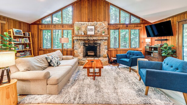 A cozy living room with a stone fireplace, wooden walls, and large windows. - Creek Access & Deck: Family Pocono Pines Cottage (Pocono Pines)