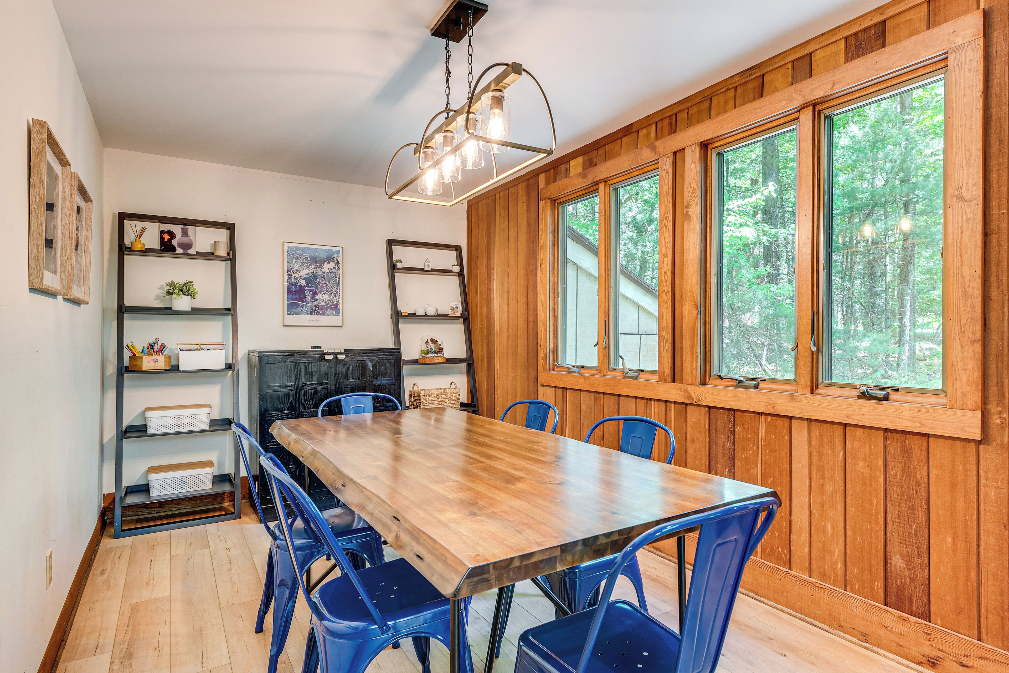 A dining area with a wooden table and blue chairs, surrounded by shelves with various items and a large window with a view of trees.