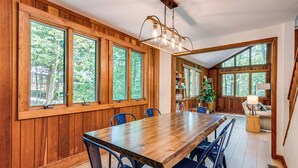 A dining area with a wooden table and chairs, large windows with a view of trees, and a ceiling light fixture. - Creek Access & Deck: Family Pocono Pines Cottage (Pocono Pines)