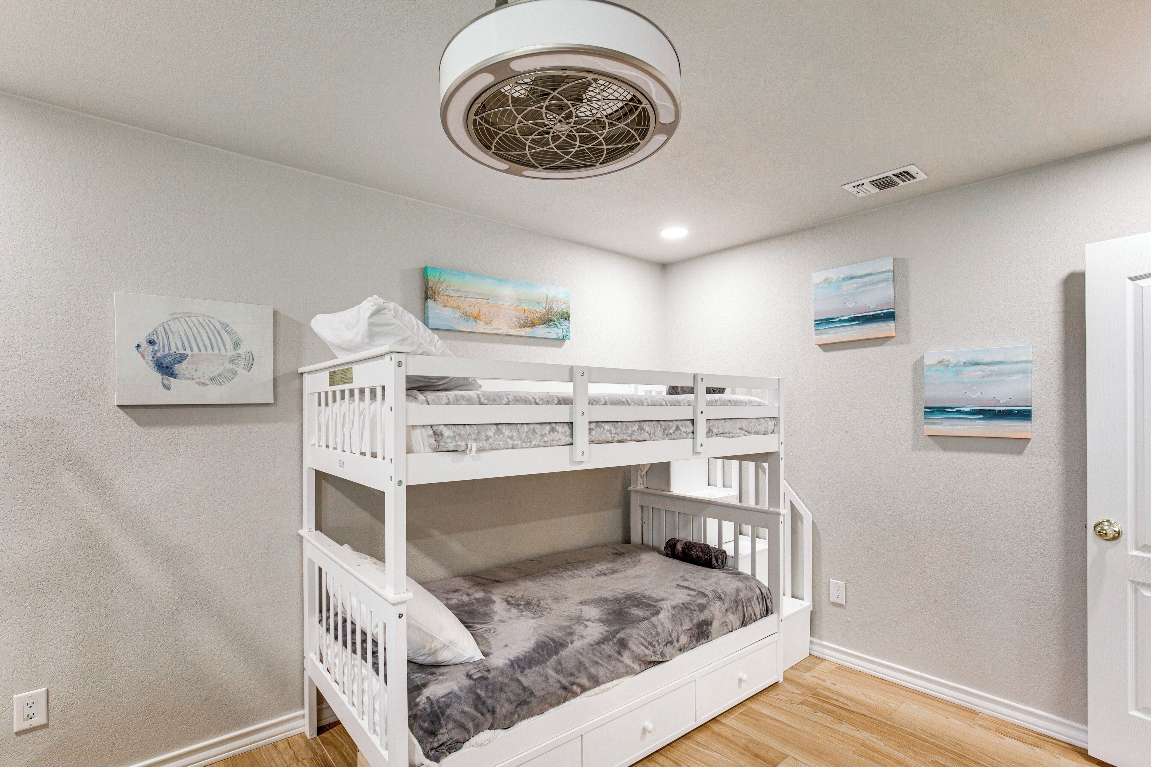 Une chambre avec des lits superposés, un ventilateur de plafond, des œuvres d’art accrochées aux murs et une porte sur le côté droit.