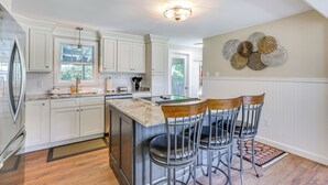 A kitchen with white cabinets, a stainless steel refrigerator, a marble countertop, and a bar area with two stools. - Old Saybrook Home w/ Screened Porch & Ocean View! (Old Saybrook)
