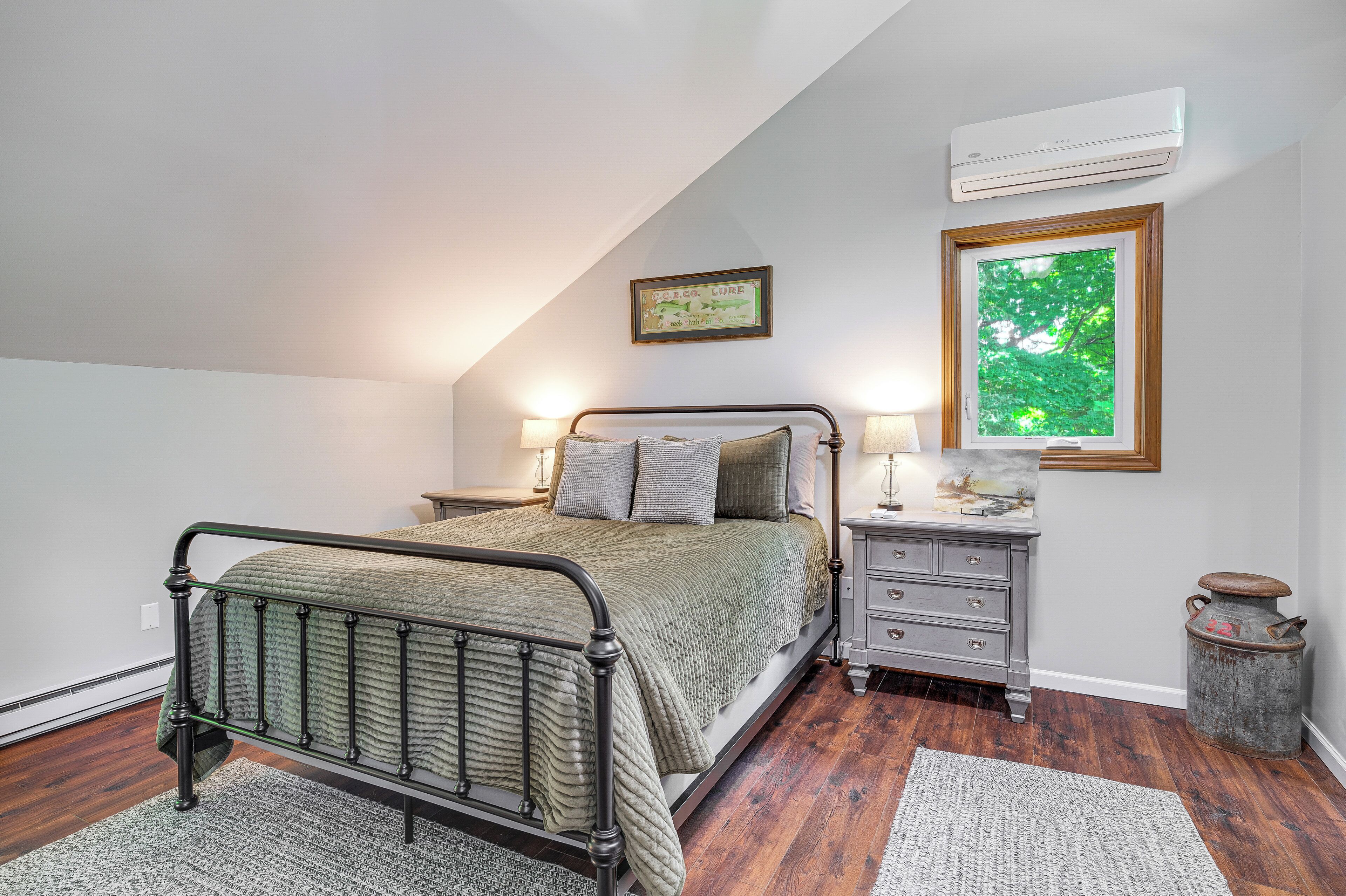 A bedroom with a bed, nightstands, a lamp, a picture frame, and a window with a view of trees.