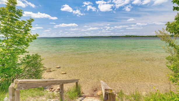 A wooden deck leading to a clear lake with a distant shoreline. - Alden Home w/ Fire Pit: Steps to Torch Lake! (Alden)