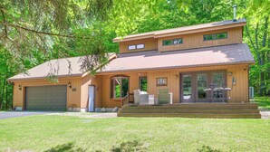 A two-story house with a garage, a covered porch with outdoor furniture, and a well-maintained lawn. - Alden Home w/ Fire Pit: Steps to Torch Lake! (Alden)