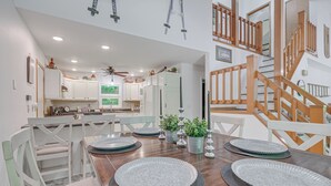 A modern kitchen with white cabinets, a wooden dining table set with plates, and a staircase with wooden railings. - Alden Home w/ Fire Pit: Steps to Torch Lake! (Alden)