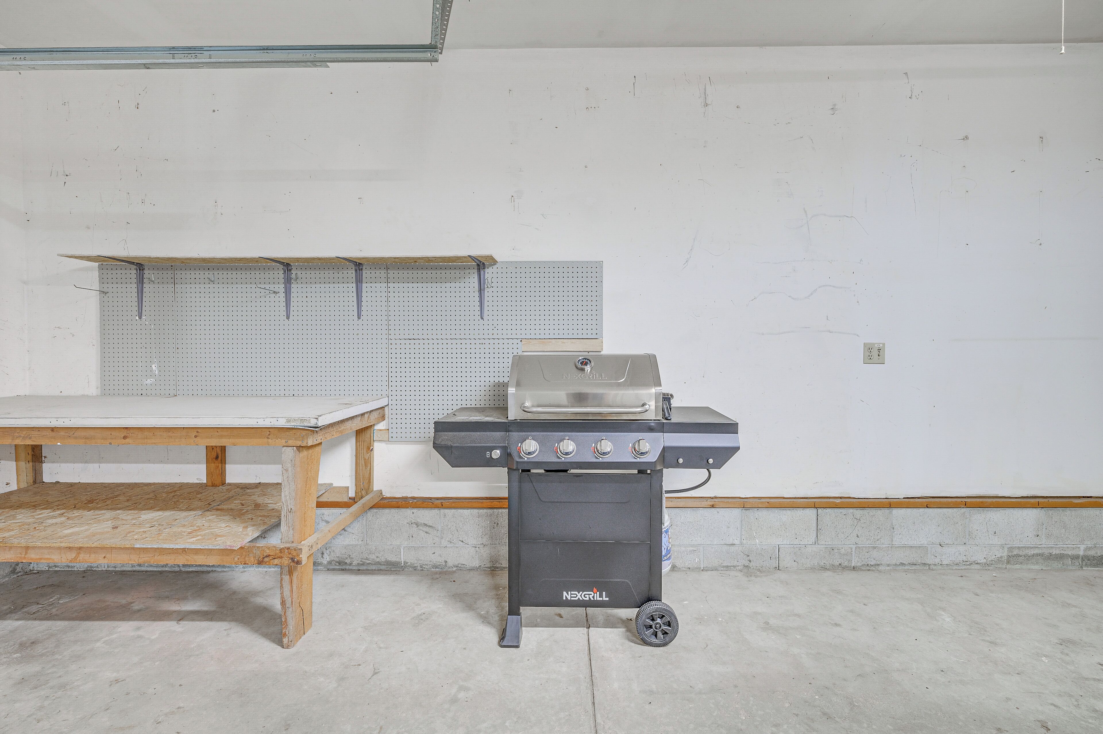 En portabel NEXGRILL-grill placerad i ett rum med en väggpanel med hål, ett träbord och ett betonggolv.