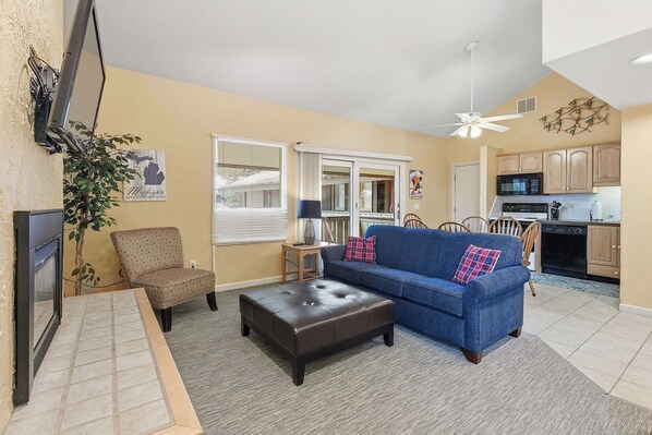 A living room with a blue sofa, a brown armchair, and a leather ottoman. There is a television mounted on the wall, a plant, and a window with blinds.