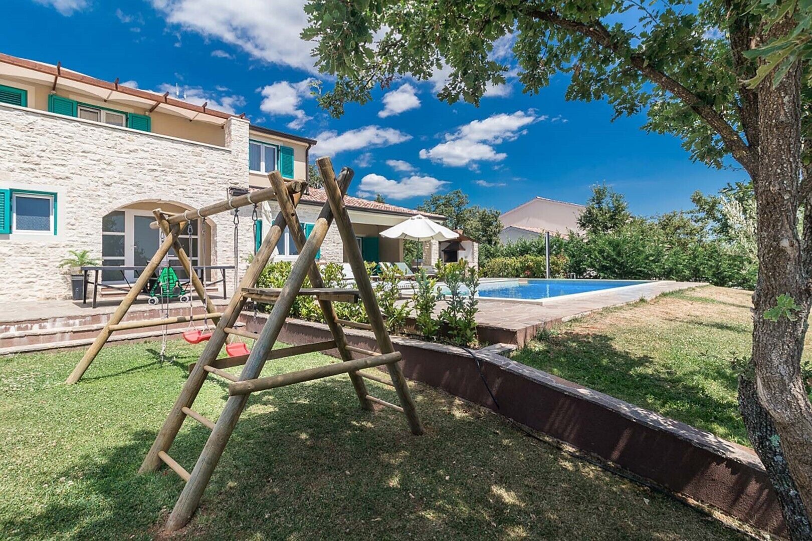 A wooden swing set in a backyard with a swimming pool and a stone building in the background.