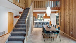 Un interior espacioso con una escalera que conduce a una zona superior tipo altiloque, un comedor con una mesa larga y sillas, y paredes de madera.