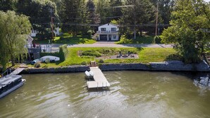 A house with a dock extending into a calm body of water, surrounded by lush greenery and a well-maintained lawn. - Chirping Pines: Modern Lake Home on Cayuga Lake (Seneca Falls)