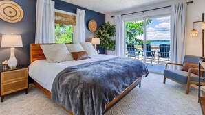 A bedroom with a large bed, a nightstand, a lamp, a window with curtains, and a view of the outdoors. - Chirping Pines: Modern Lake Home on Cayuga Lake (Seneca Falls)