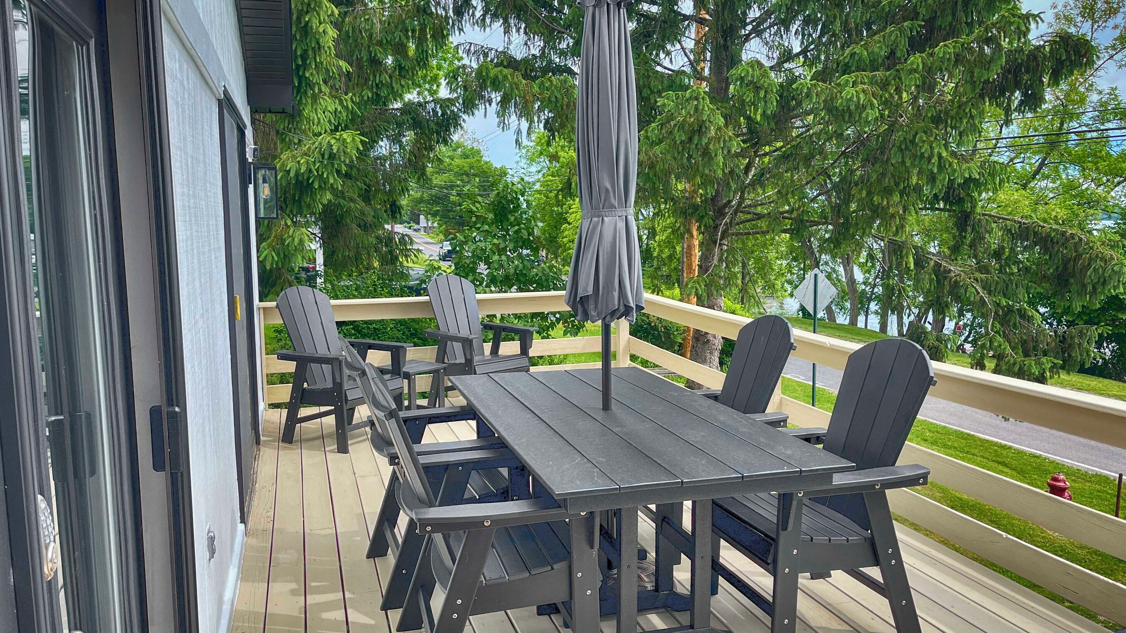 A deck with a table and chairs, surrounded by a railing, overlooking a road and greenery.