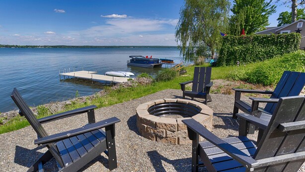 A lakeside deck with Adirondack chairs, a fire pit, and docked boats. - Chirping Pines: Modern Lake Home on Cayuga Lake (Seneca Falls)