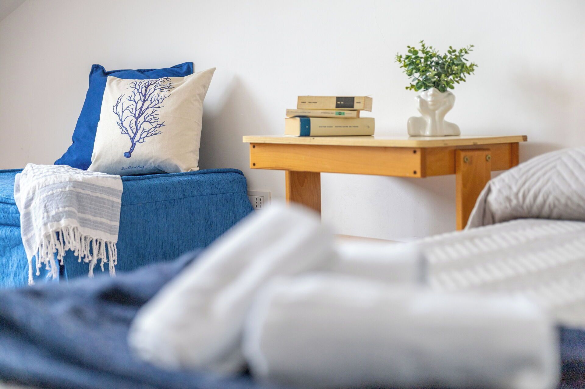 A cozy room with a blue sofa, a white pillow with a tree design, a small wooden table with books and a vase, and a white blanket on the floor.