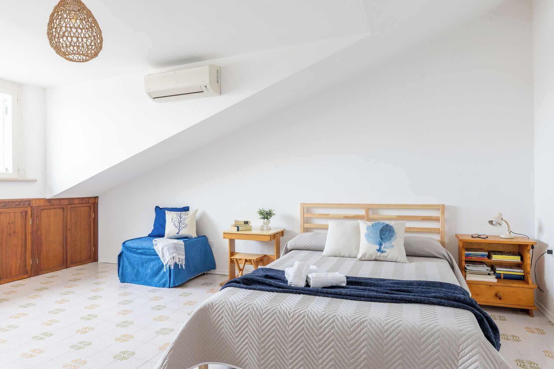 A bedroom with a sloped ceiling, a bed with a wooden headboard, a blue chair, a bedside table with books, and a wall-mounted air conditioner.
