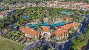 An aerial view of a residential area with a central swimming pool surrounded by houses, palm trees, and a curved road.