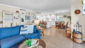 A modern living room with a blue sofa, a dining area with wooden chairs, a kitchen with white cabinets, and a large window. - ☀️Pelican Pointe 4C- Beachfront Views from Balcony☀️ (Indian Shores)