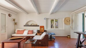 A living room with a sofa, coffee table, and a dining table. There is a window with a view of trees and a door leading outside.
