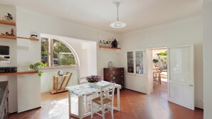 A kitchen with a dining table, chairs, and a window. There is a cabinet with drawers and a shelf with decorative items.
