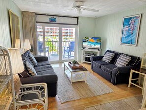A living room with a sofa, armchair, coffee table, TV, and balcony doors. - Oceanview Midtown Condo with Pool! Seaside 66 - #202 (Ocean City)