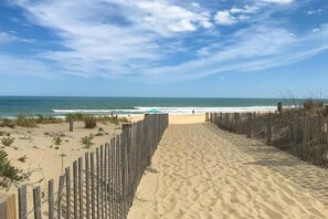 A sandy beach path leading to the ocean with a wooden fence on one side and a clear blue sky above.