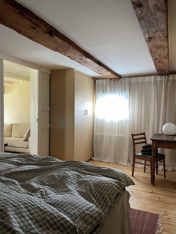 A bedroom with a large bed, wooden beams on the ceiling, a desk with a chair, and a window with curtains.