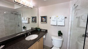 Een moderne badkamer met een granieten blad, een witte wastafel, een toilet en een glazen douchecabine. Handdoeken hangen aan een rek en er hangt een afbeelding in een lijst aan de muur.