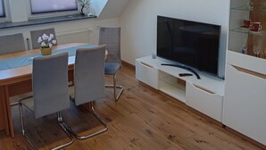 Moderni, hyvin valaistu huone, jossa on ruokailutila, suuri litteänäyttöinen televisio ja kalteva katto.