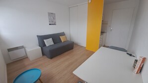 Een compacte, moderne kamer met een grijze bank, een blauwe fauteuil, een witte tafel en een geel accentmuur.