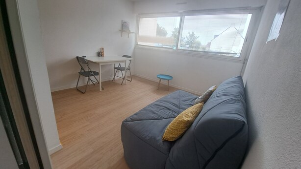 Een knus woongedeelte met een grijze bank, een tafeltje, twee stoelen en een raam met zonwering.