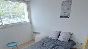 Een slaapkamer met een bed, een klein tafeltje en een stoel. Er is een raam met zonwering, een poster aan de muur en een radiator.