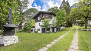 Un chalet à deux étages, avec un chemin de pierre menant à l’entrée, entouré d’une végétation luxuriante et de montagnes.