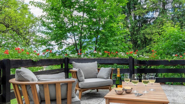 Un espace extérieur aménagé avec deux fauteuils gris rembourrés et une table en bois, le tout mis en valeur par un décor de verdure luxuriante et une clôture en bois.