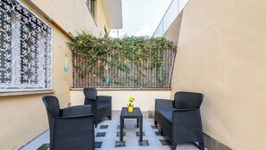 Eine kleine Außenveranda mit Korbmöbeln und einem Tisch, ein Fenster mit dekorativen Metallgitter und eine mit Kletterpflanzen bewachsene Wand.