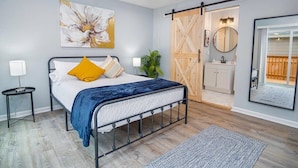 A bedroom with a bed, a nightstand, a wall-mounted mirror, a sliding barn door closet, and a large flower painting. - Spacious Delightful 4 BR/ 3 BA Close to Downtown (San Antonio)